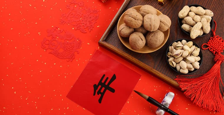 Red background with Chinese New Year decorations, including paper-cuts, calligraphy with the character "年", walnuts, peanuts, pistachios, and a red knot.