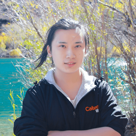 一位留长发的年轻人,身穿印有“Caltech 18”字样的黑色夹克,站在户外碧绿的湖水和树木前微笑。