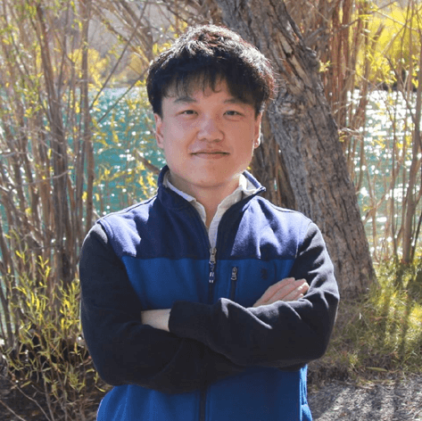 一位年轻亚洲男子,双臂交叉,面带微笑,身穿蓝黑夹克,背景是树木和波光粼粼的水面。