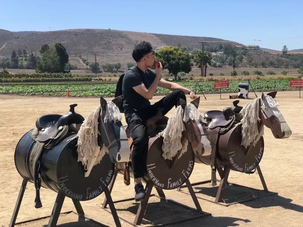 A person sits on a barrel-horse ride at Underwood Family Farms, holding a red object. Other barrel-horses, fields, and hills are in the background.