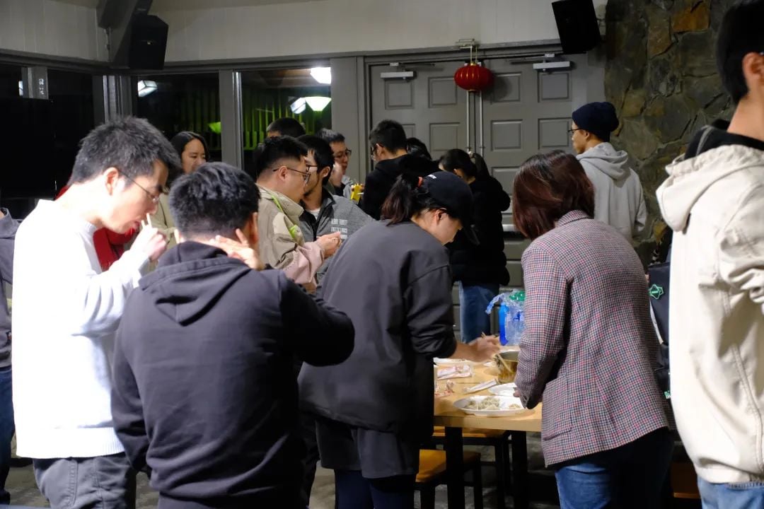 室内聚会,人们围桌而立,有人在吃饺子,背景有红灯笼。