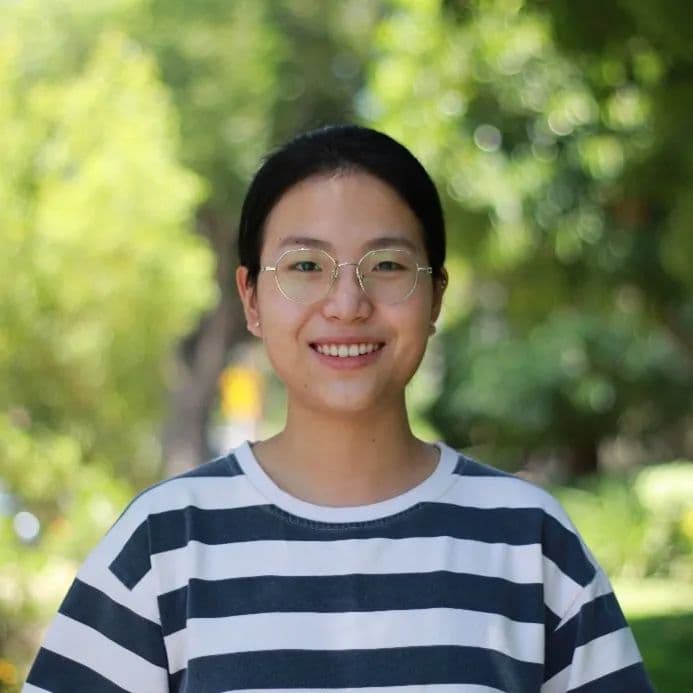 一位戴眼镜、穿条纹衫的年轻东亚女性,在模糊的绿色户外背景前微笑。