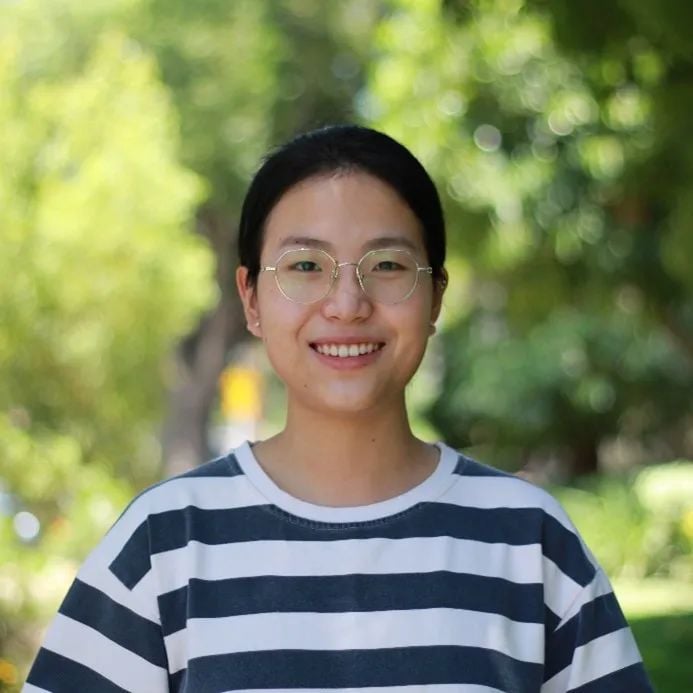 一位戴眼镜、穿条纹衫的年轻东亚女性,在模糊的绿色户外背景前微笑。