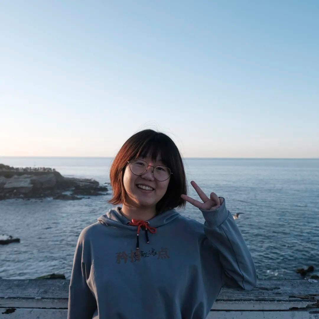 一位戴眼镜的年轻女子在海边微笑,比出V字手势。