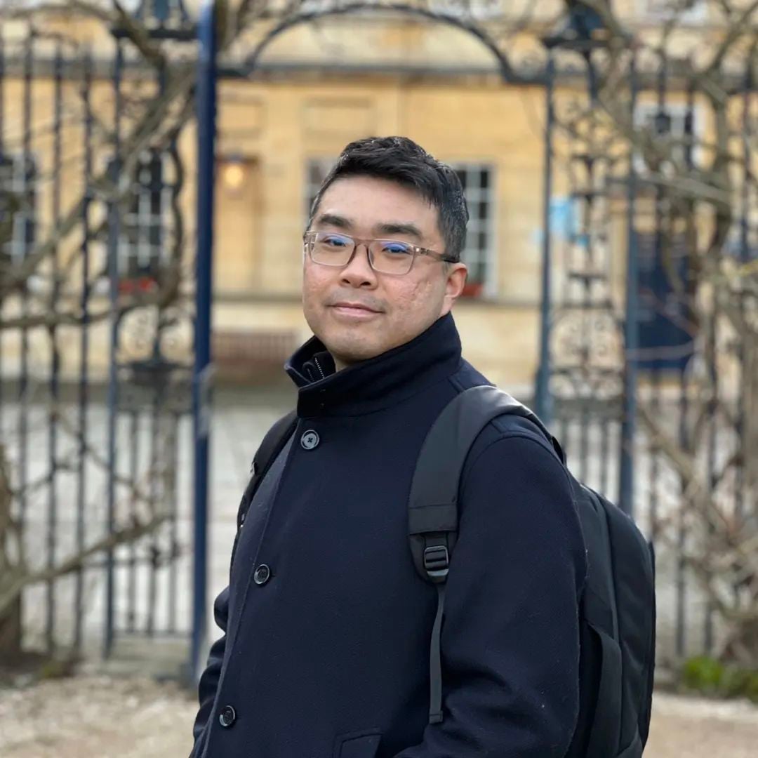 戴眼镜、穿深色外套和背包的男子,站在华丽大门和建筑前,看向镜头。