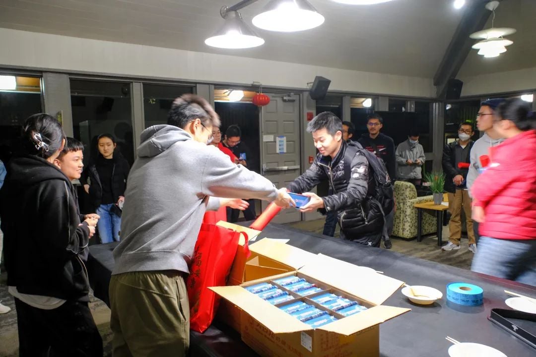 活动中,一人将蓝色物品递给另一人,桌上有装满物品的盒子和红色袋子。