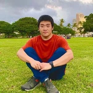 年轻男子盘腿坐在草地上,背景是树木和建筑。