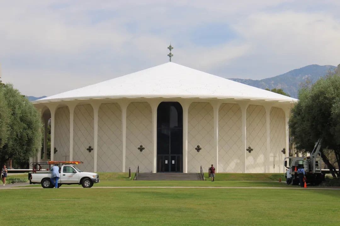 A round, light-colored building with a white conical roof on a green lawn. Trucks are parked in front, with mountains in the background.