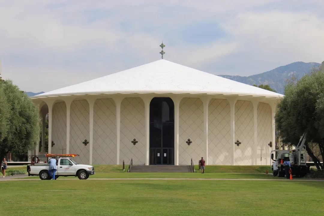 圆形浅色建筑,白色锥形屋顶,前方有草坪和车辆,背景是山脉。