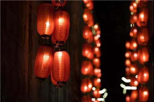 Glowing red lanterns hang in a dark setting, some in focus on the left, many blurred on the right, creating a warm, festive scene.