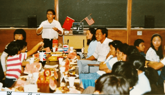 人们围坐桌旁,桌上摆满食物饮料;一人站立发言,旁有中美两国国旗。