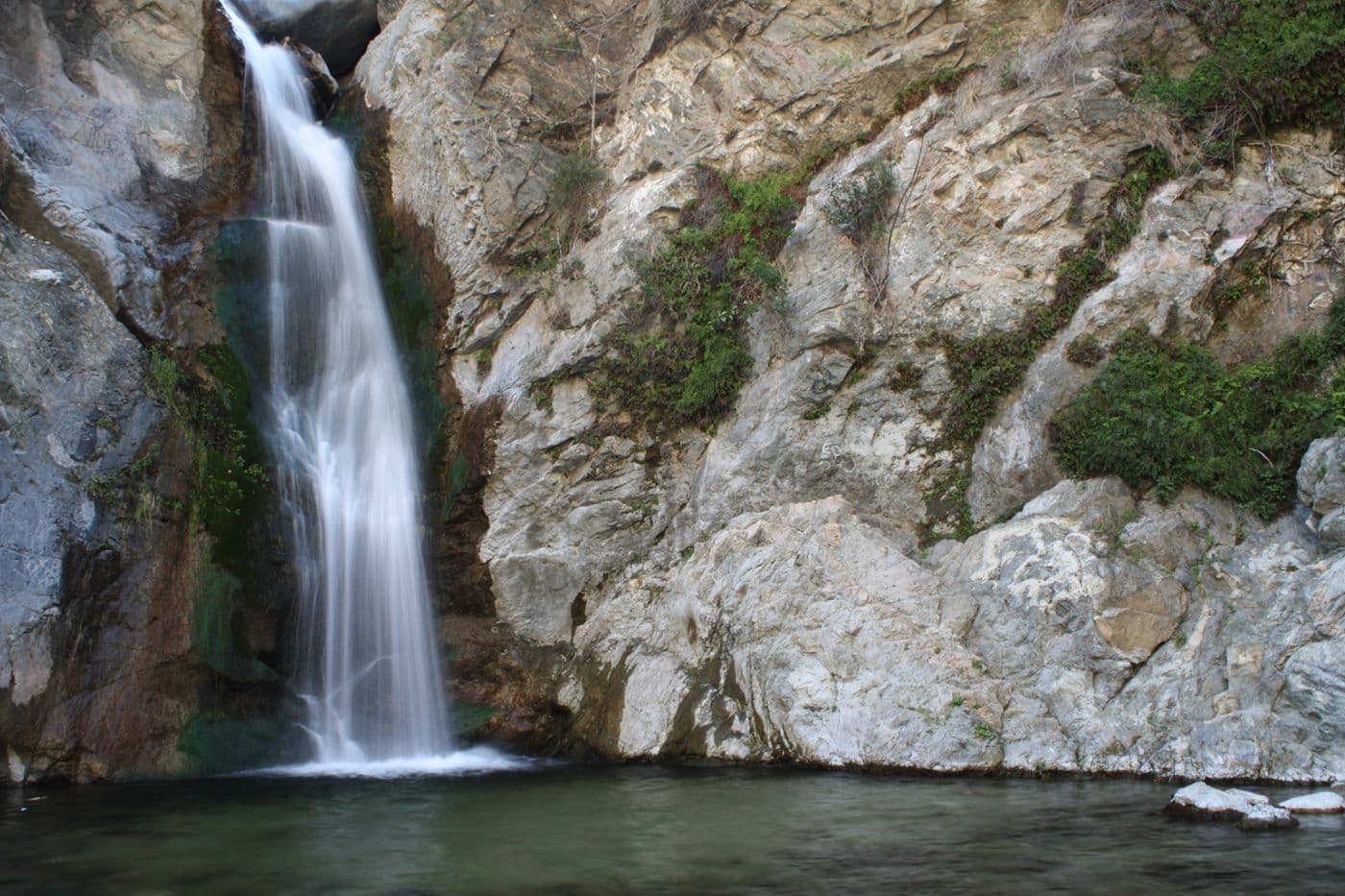 A long exposure shot of a waterfall cascading down a rocky cliff face into a calm pool of water, with green vegetation growing on the rocks.