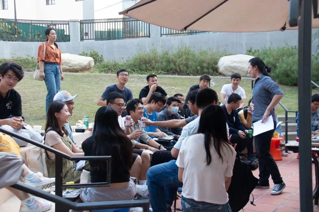 一群年轻人在户外聚会,有人坐在草地和台阶上,有人站着,在遮阳伞下交谈。