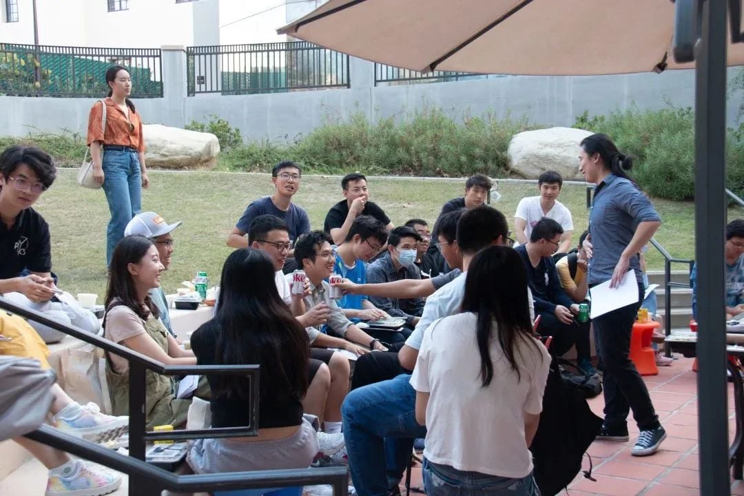 一群年轻人在户外聚会,有人坐在草地和台阶上,有人站着,在遮阳伞下交谈。