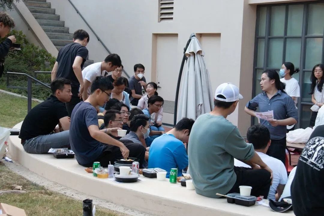 一群年轻人在户外聚会,部分人坐在矮墙上用餐,一人站立向大家讲话。
