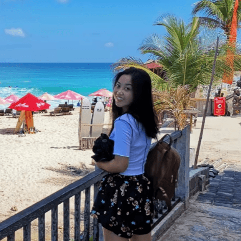 一位年轻女子在海滩边,面带微笑地看向镜头,背景是蓝天碧海和沙滩伞。