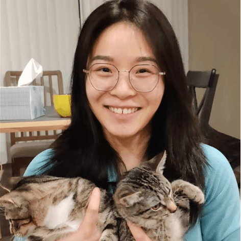 一位戴眼镜的年轻女子抱着两只虎斑猫,面带微笑。背景有纸巾盒。