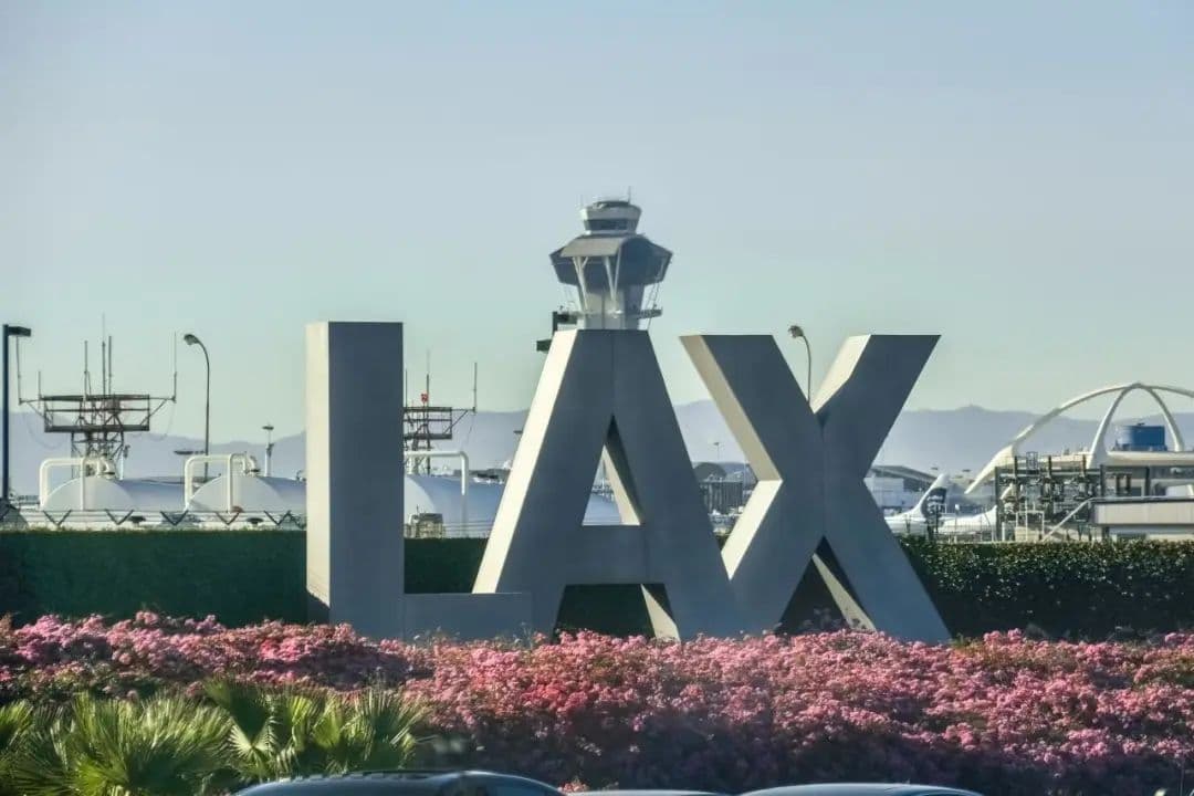 Large "LAX" sign with the airport control tower behind it, surrounded by pink flowers and green bushes in the foreground, under a clear sky.