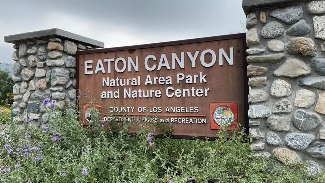 一个棕色标牌,上面写着“EATON CANYON自然区公园和自然中心”,两侧是石柱,前景有紫色花朵和绿色植物。