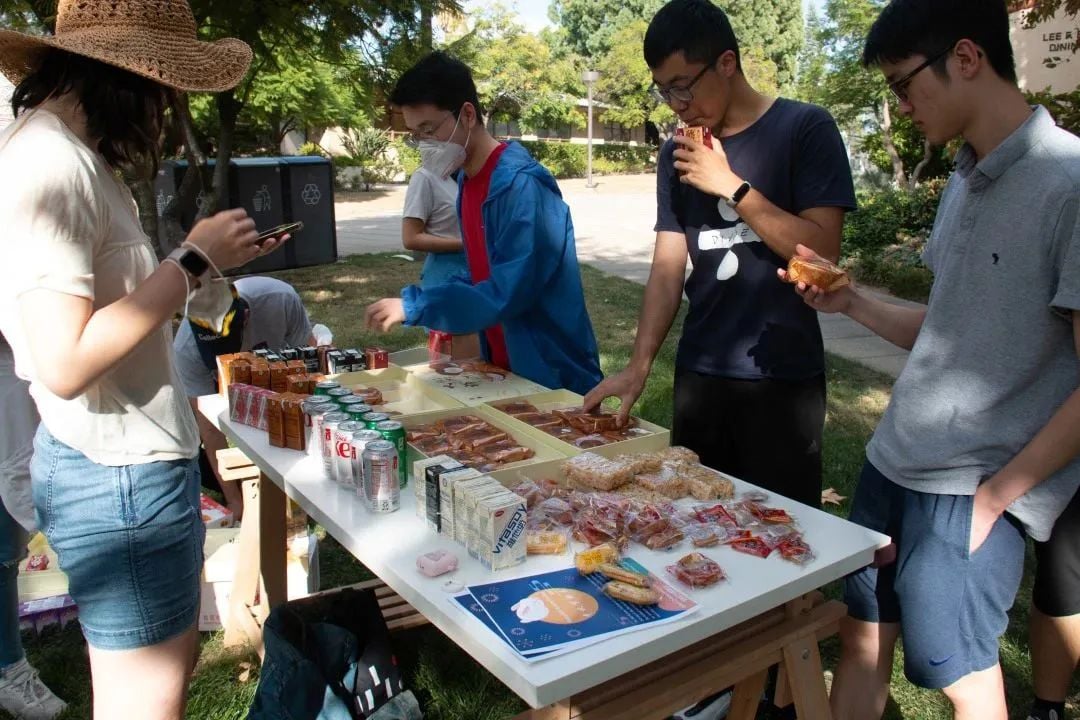 学生们在户外小吃摊前,桌上摆放着饮料、糕点和零食,几名学生正在挑选或品尝。